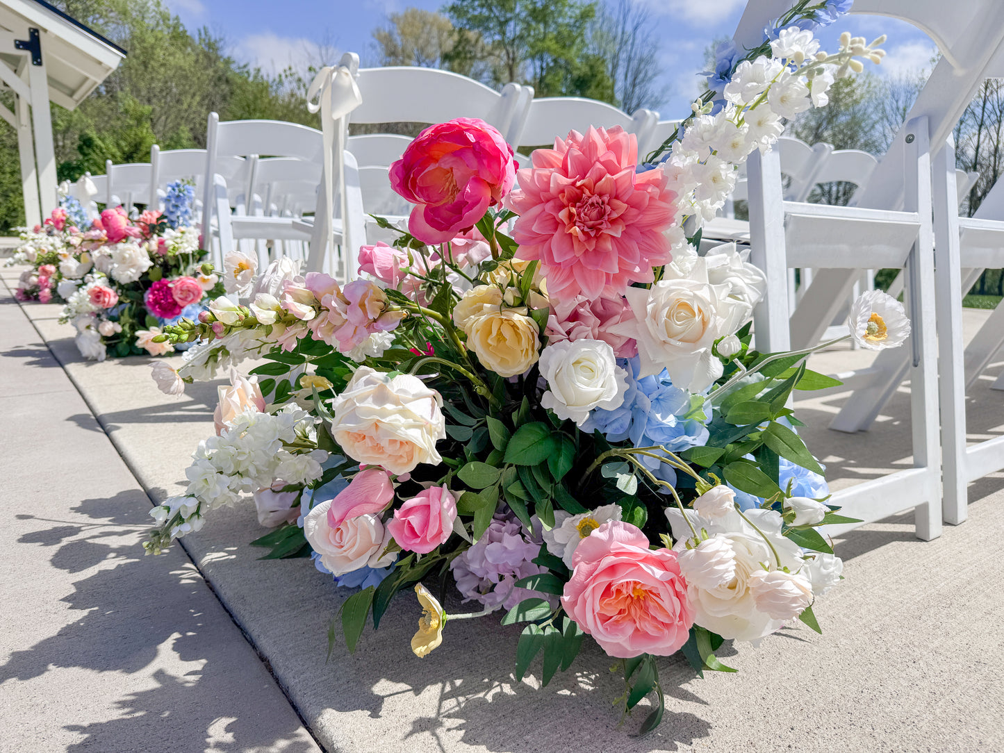 A large floral arrangement of pink, white, yellow, and blue flowers with green leaves, placed along the aisle in front of white folding chairs outdoors.