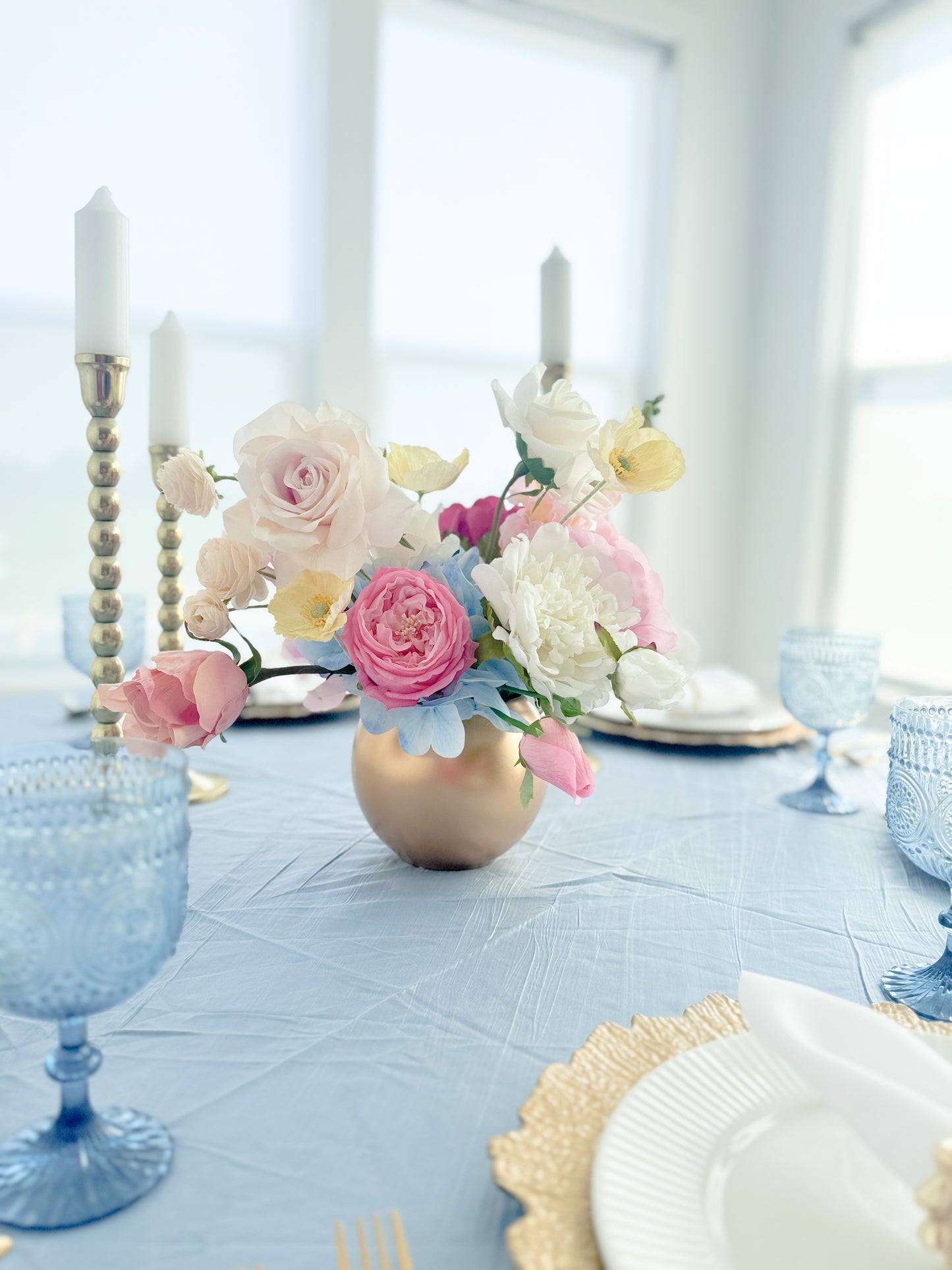 A pastel floral centerpiece in a metallic gold vase on a light blue tablecloth, styled with blue glass goblets, white plates, and candlesticks in a bright dining room.