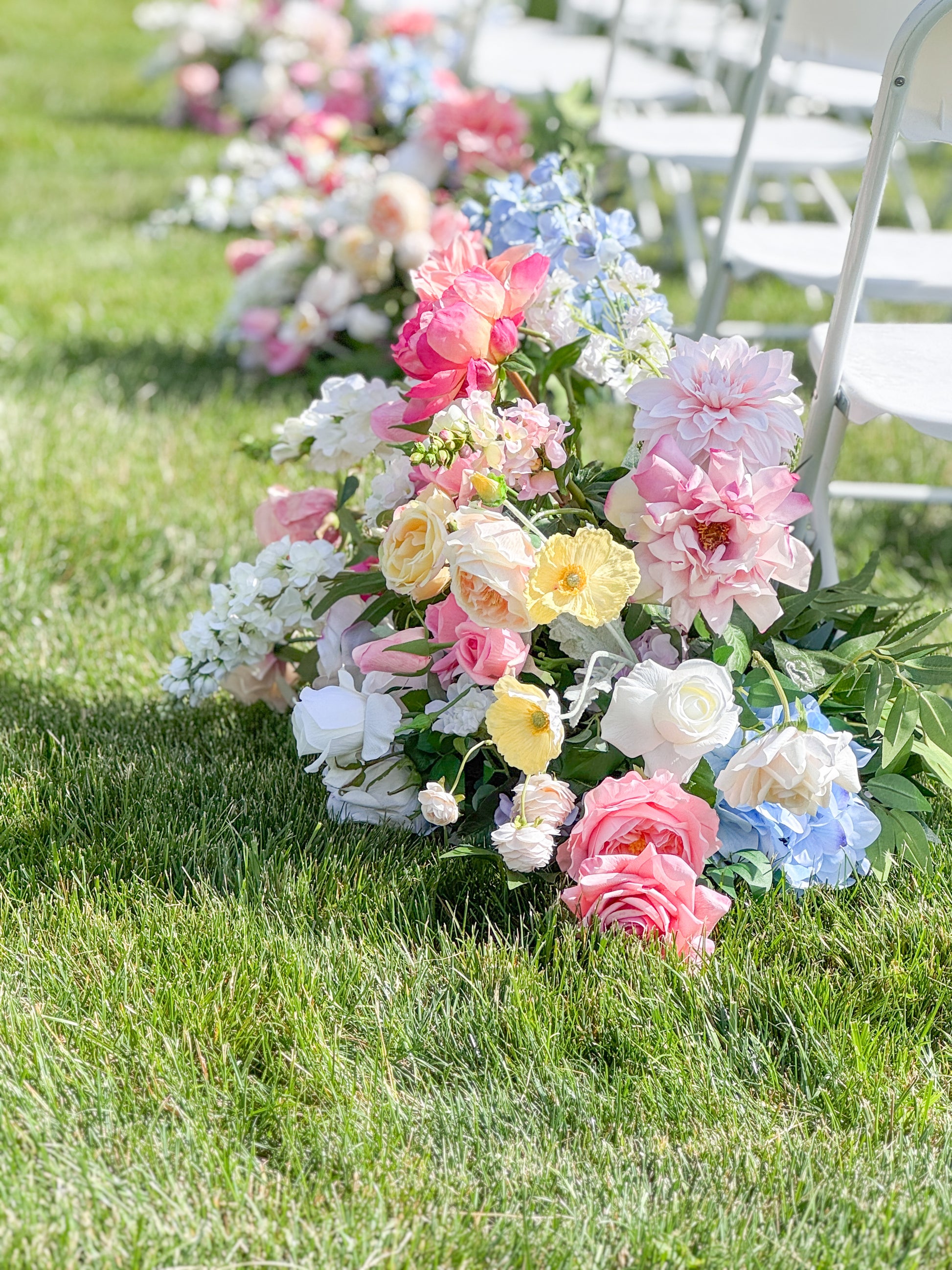 A long floral arrangement of pastel pink, white, yellow, and light blue flowers placed along the edge of a grassy outdoor area beside white folding chairs.