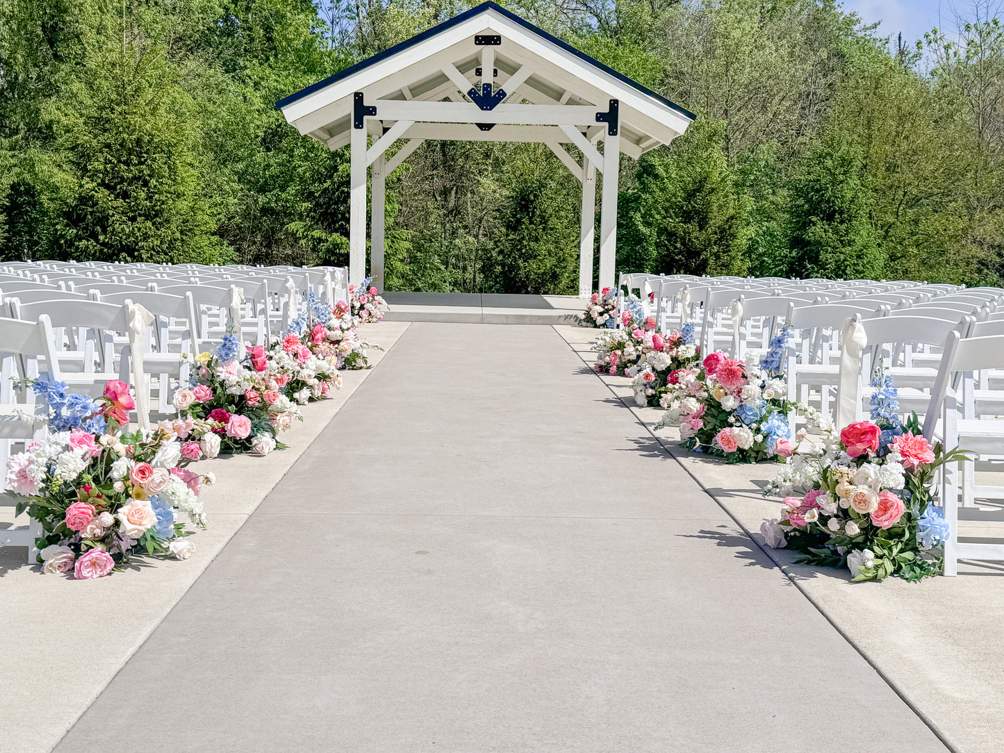 Outdoor wedding ceremony setup with a white wooden pavilion at the front, rows of white chairs on both sides, and floral arrangements in pink, blue, and white along the aisle and seat edges, with green trees in the background.