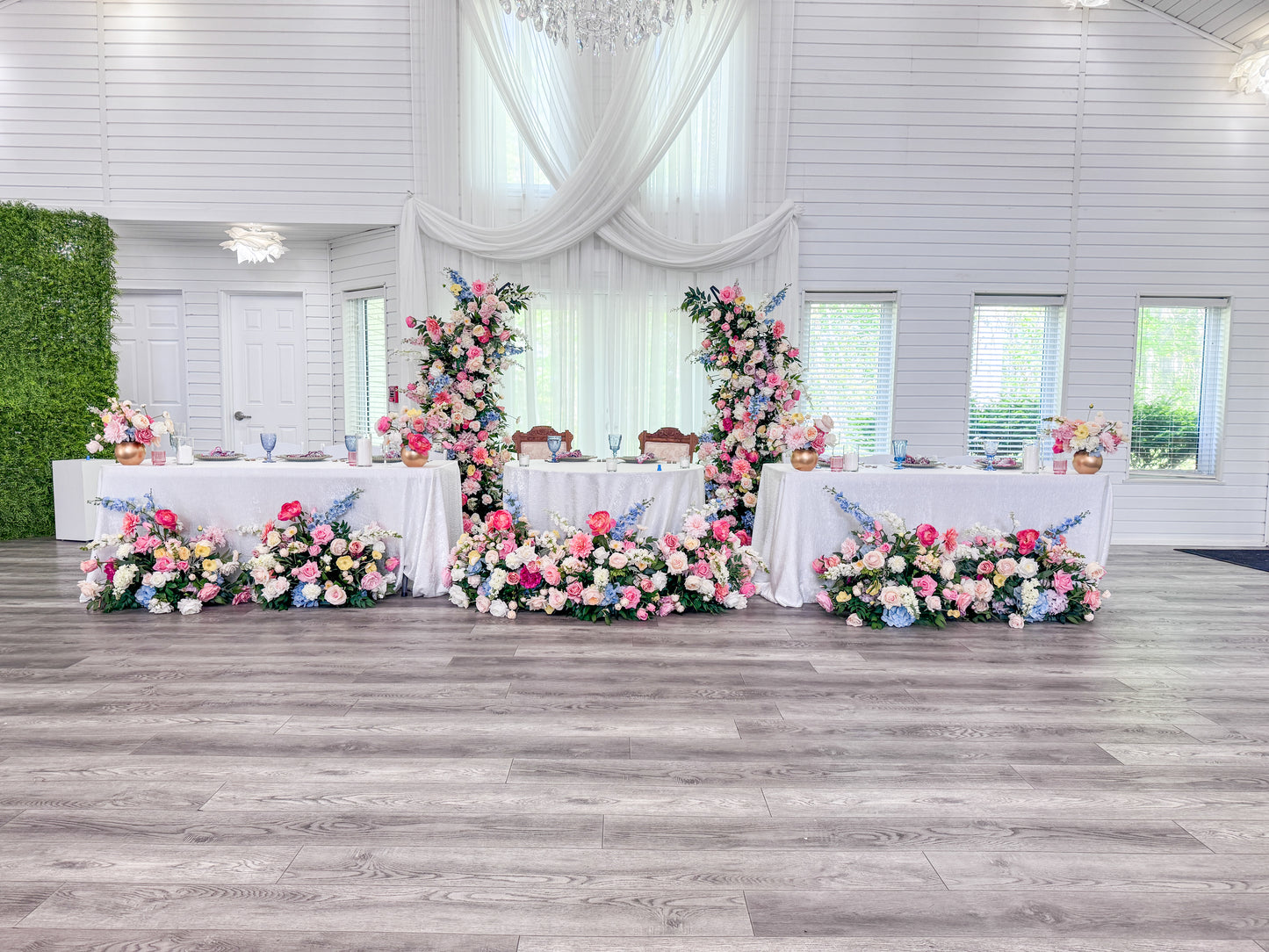 A decorated event space with three long white tables covered in pink, blue, and white floral arrangements, set on a gray wood floor in a bright white hall with draped curtains and a green wall panel.