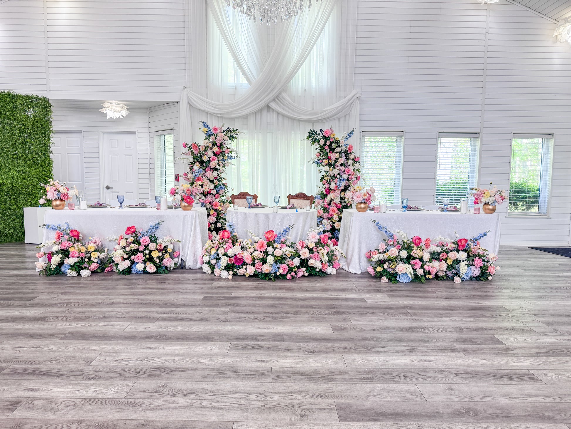 A decorated event space with three long white tables covered in pink, blue, and white floral arrangements, set on a gray wood floor in a bright white hall with draped curtains and a green wall panel.