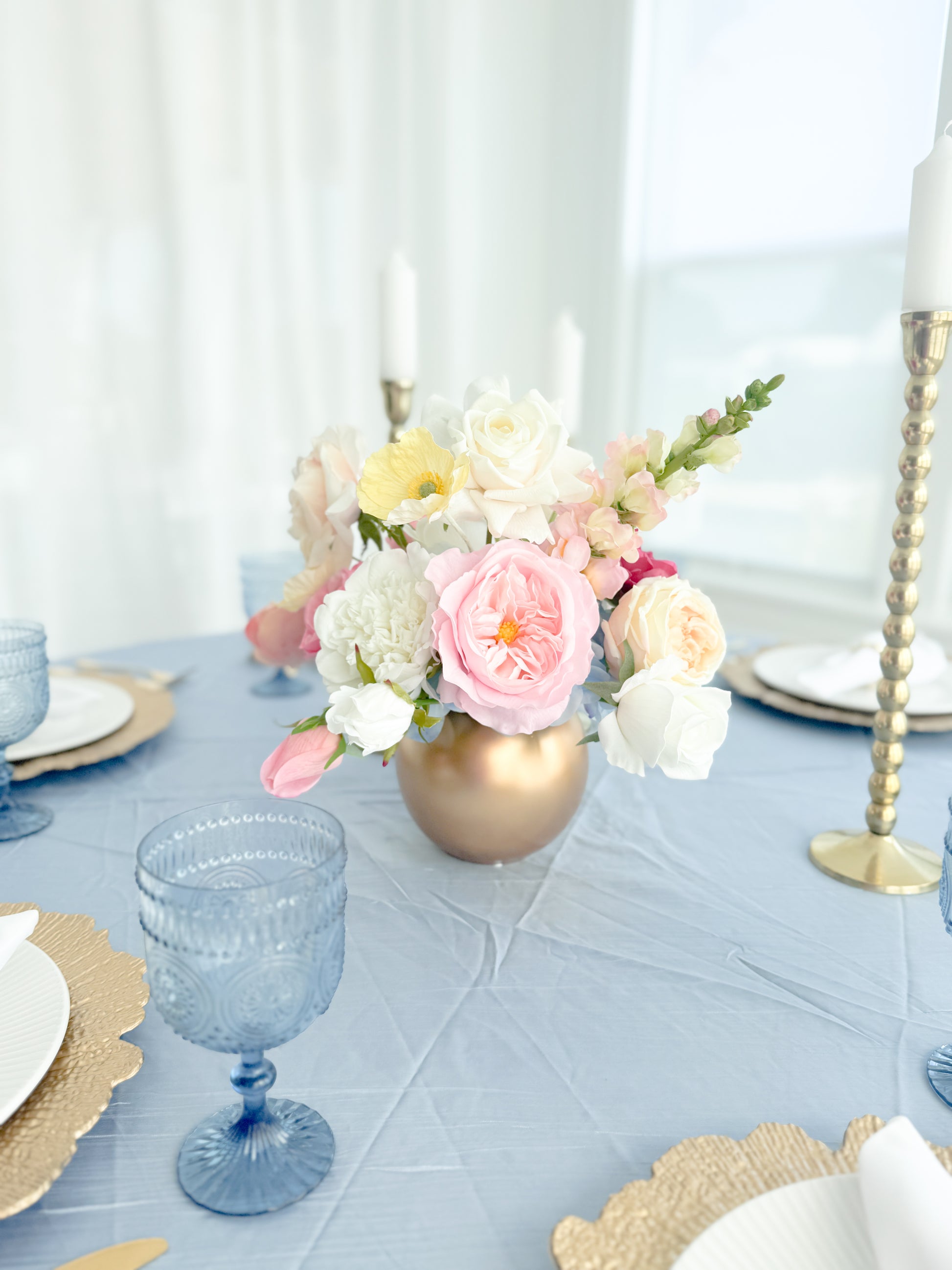 A floral centerpiece arrangement in a metallic gold vase on a light blue tablecloth, surrounded by blue and gold glassware and plates in a bright dining setting.