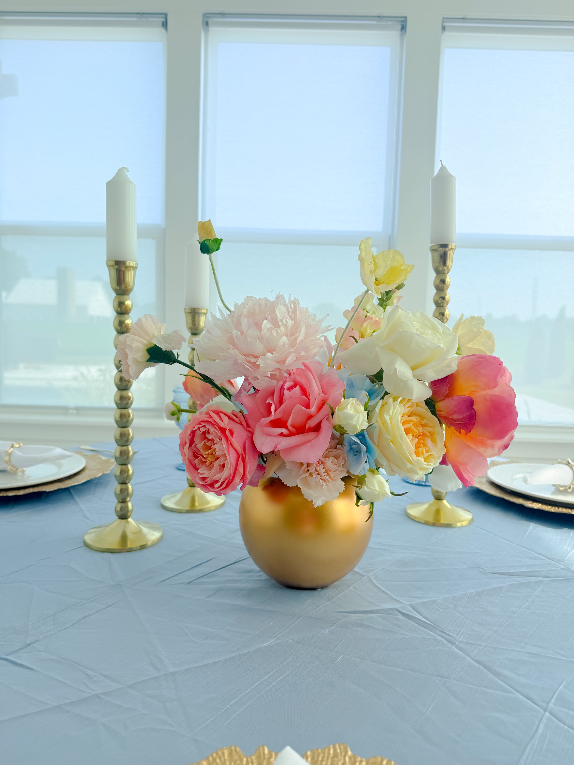 A floral centerpiece arrangement in a round gold vase on a light blue tablecloth, with two gold candlesticks and plates set on either side near large windows.
